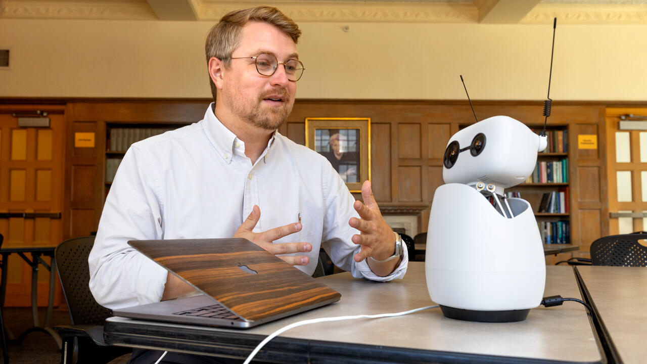 Scott Seidenberger at a library table, talking with Hopper, a small white robot with round camera-lens eyes.